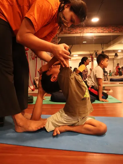 I provide hands-on assistance to help this young student safely deepen his Eka Pada Rajakapotasana (One-Legged King Pigeon Pose), ensuring correct alignment.