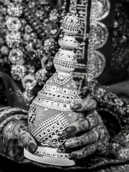 A detailed black and white shot of a bride's hands, adorned with henna, holding a traditional pot during a wedding ritual.