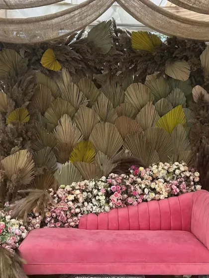 A chic boho-themed seating area for a pre-wedding event. A vibrant pink chaise lounge is set against a backdrop of dried palm leaves and pampas grass, surrounded by fresh flowers.