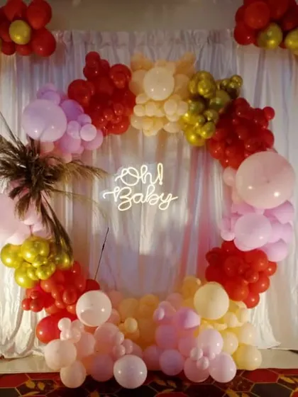 An "Oh Baby" baby shower decoration with a balloon ring in warm tones of red, pink, and gold, set against a white curtain backdrop.
