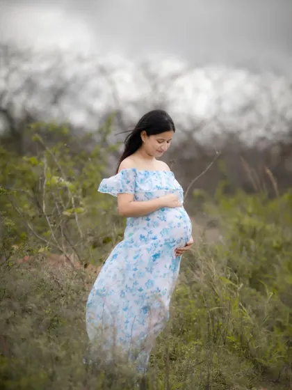 A serene outdoor maternity portrait. The mom-to-be is standing in a field of green, wearing a beautiful blue and white floral dress, looking peacefully at her baby bump.