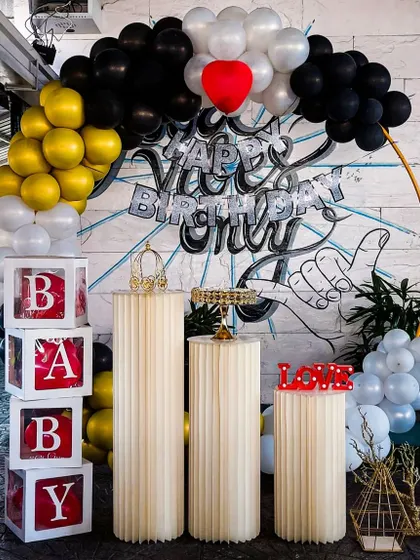 For a cool and urban vibe, we created this setup featuring a black, gold, and white balloon arch against a graffiti art wall. The arrangement includes 'BABY' blocks and fluted pedestals, blending modern decor with street art style.