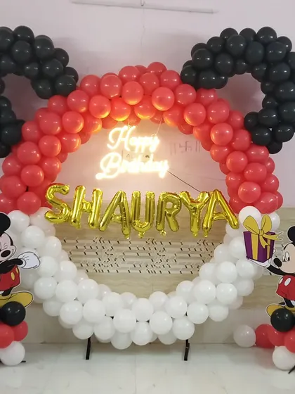 A unique Mickey Mouse head-shaped balloon ring backdrop with a neon sign and character cutouts, perfect for photos at a birthday party.
