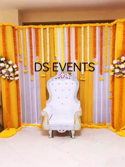 A simple and elegant backdrop for a Haldi or small ceremony. The stage is decorated with yellow and white drapes and hanging marigold garlands, with a white throne-style chair as the centerpiece.