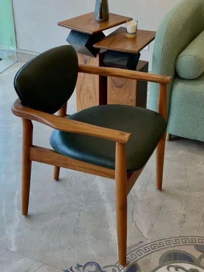 The "Catwalk" chair features a sleek teak wood frame and a floating seat in deep green leatherette. Its elegant, mid-century inspired design makes it a versatile accent chair for any modern living space.