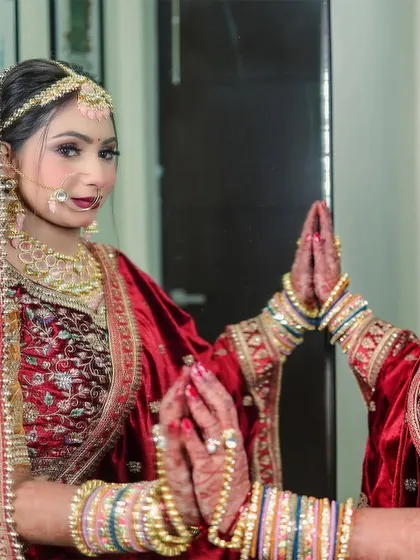 The bride looks into the mirror, her hands joined in a reflective pose. This image captures the beauty of her traditional red velvet lehenga and the quiet moments of anticipation before the ceremony.