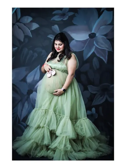A sweet portrait of a mom-to-be holding tiny baby shoes against her bump. She is wearing a green ruffled gown against a dark floral backdrop.
