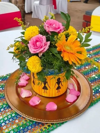 A close-up of a table centerpiece for a Mehendi party. The bright bouquet of pink and yellow flowers is placed in a hand-painted pot with traditional Warli art, sitting on a colorful sequined table runner.