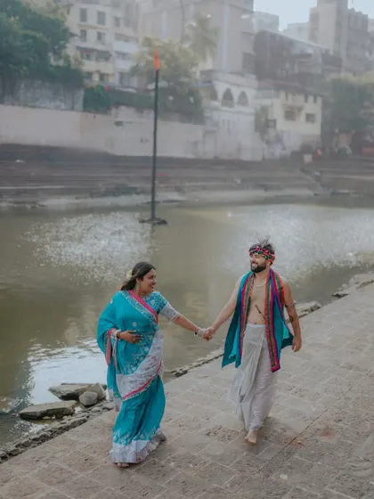 Walking along the ghats, hand in hand. This wide shot captures the couple in their beautiful traditional attire against the backdrop of the river and old city buildings.
