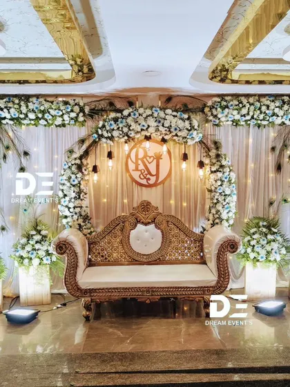 An elegant stage setup combining a circular floral arch with delicate peacock feather accents. The design uses a mix of blue and white flowers, complemented by a custom monogram and a classic gold and cream sofa.