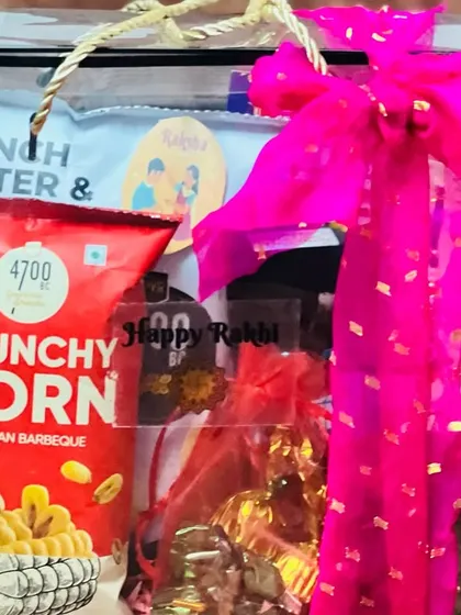 A clear box hamper for Rakhi, filled with crunchy corn snacks and chocolates, tied with a bright pink bow.