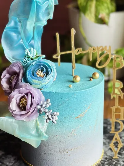 Elegance in every bite. This blue and lilac ombre cake is a dreamy blend of delicate hues, topped with sugar flowers and an ethereal sail for a magical birthday celebration.