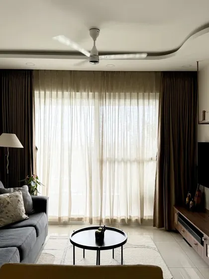 A modern living room with a comfortable grey sofa, sheer curtains, and a curved false ceiling detail.