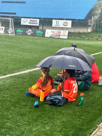 Players waiting on the sidelines under umbrellas during a rainy match day.