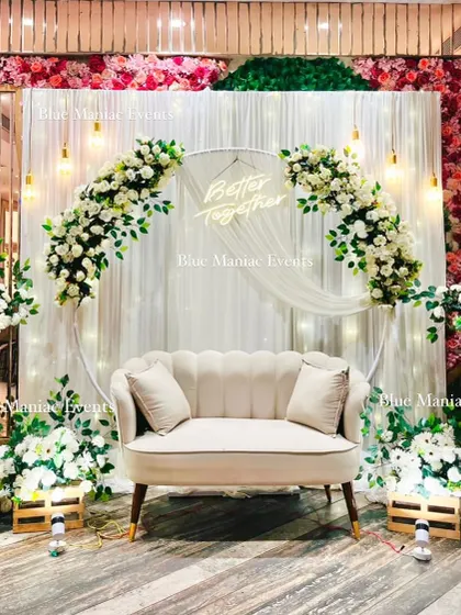For a sophisticated and timeless celebration, we designed this beautiful white-themed Roka setup. It features a circular arch with white and pink floral arrangements, a 'Better Together' neon sign, and elegant lighting against a backdrop of red and green flower walls.