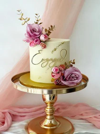 A classic engagement cake with a smooth buttercream finish and gold leaf details. The design is completed with an arrangement of purple and pink silk roses.