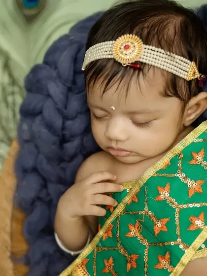 A delicate close-up of baby Radha sleeping, showing the details of her traditional headband and outfit.
