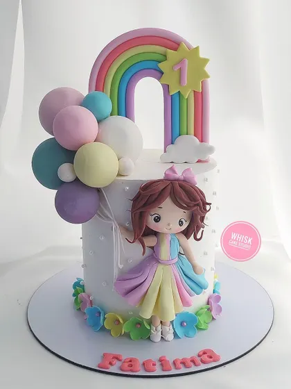 A beautiful and colorful first birthday cake. This design features a handcrafted doll, a pastel rainbow, and a cluster of colorful balloons.