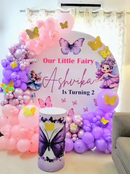 A fairy and butterfly themed second birthday decoration for Ashvika. The setup features a custom backdrop with fairies, a purple and pink balloon arch, and butterfly cutouts.