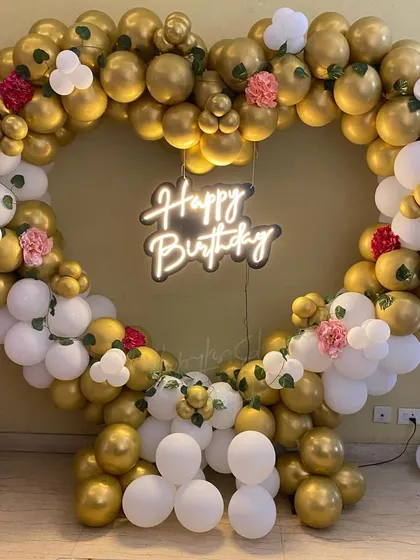 A stunning heart-shaped birthday balloon arrangement in white and gold, with floral accents and a neon sign.
