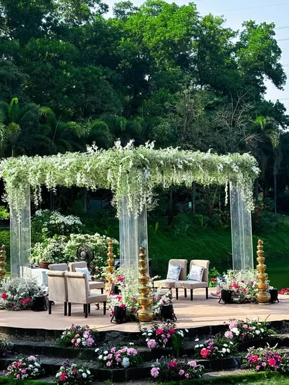 The lakeside mandap design, featuring acrylic pillars and a ceiling of hanging white flowers, offers a breathtaking view and an atmosphere of pure elegance for the wedding ceremony.
