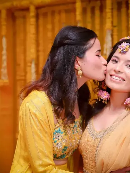A sweet moment of sisterly love at the Haldi ceremony. A gentle kiss on the cheek against a backdrop of yellow flowers makes for a beautiful, warm photo.