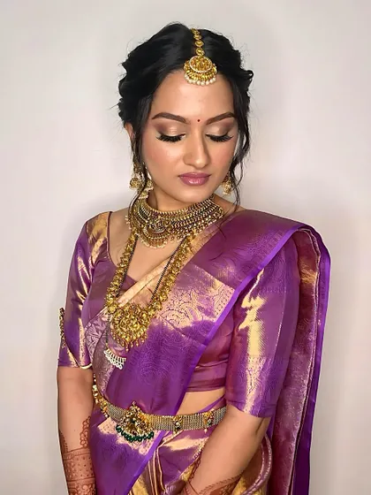 A stunning South Indian bride in a rich purple Kanjivaram saree. The makeup is pure glamour, with shimmering golden eyes, a flawless base, and intricate temple jewelry to complete the look.