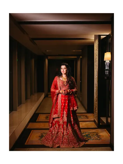 A full-length portrait of the bride in her red reception lehenga, looking away with a gentle smile. The warm, moody lighting of the hallway adds to the elegance.
