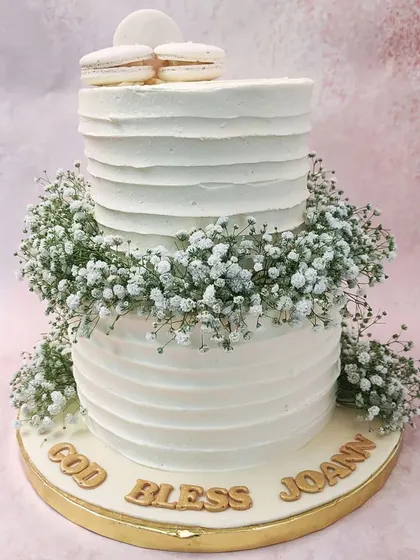We offer a blessing for a sacred milestone with this elegant two-tiered Holy Communion cake. A wreath of delicate baby's breath separates the textured white tiers, which are topped with pristine white macarons, creating a look of pure serenity.