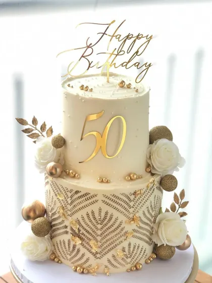 A grand two-tiered cake for a 50th birthday. The intricate gold stenciling and white roses create a look of pure luxury.