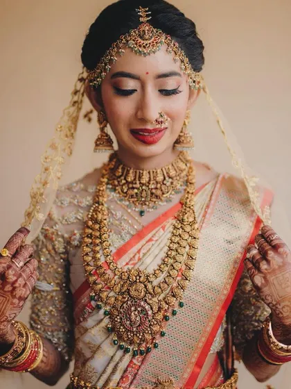 A candid moment captured. The bride's makeup, featuring a classic red lip and gold jewellery, is perfectly complemented by her happy and radiant expression.