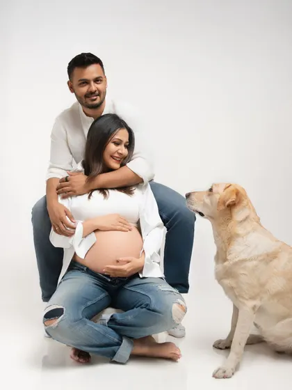 A classic maternity pose with the couple and their dog looking on. The father-to-be has his arms wrapped around the mother, creating a feeling of love and protection.