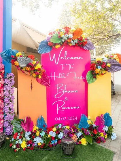 Welcome to the Haldi Fiesta. A bright pink welcome sign adorned with colorful flowers and palm leaves immediately sets a fun, tropical, and celebratory mood.