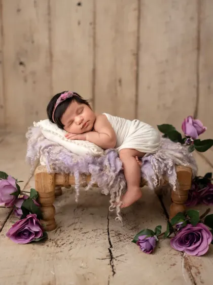 A precious newborn girl sleeping on a miniature wooden bed, surrounded by purple roses. This posed shot showcases her tiny features in a whimsical, romantic setup.