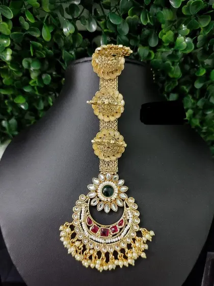 An elaborate temple-style maang tikka with multiple tiers and a crescent pendant. The red and green stones add to its traditional grandeur.