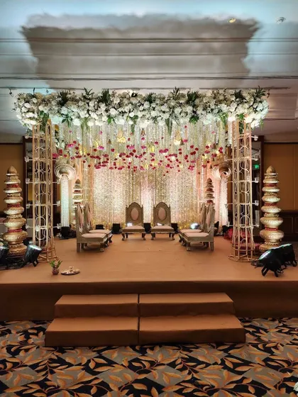 An elegant indoor mandap setup with a metal frame, adorned with a dense canopy of white flowers and hanging floral strings. The traditional stacked 'kalash' elements add a cultural touch.