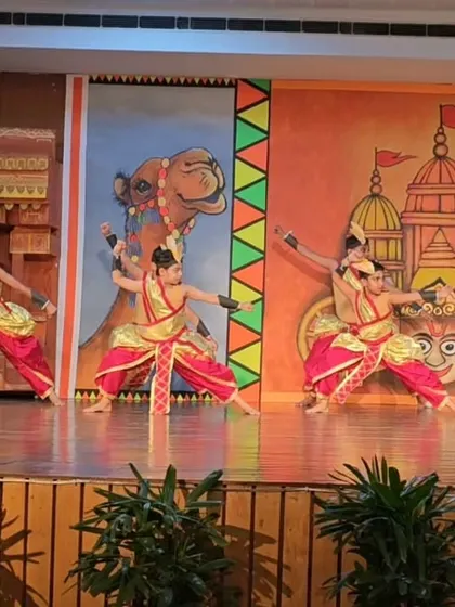 A group of young dancers in costume, performing a Chhau piece on a stage with a Rajasthani-themed backdrop.