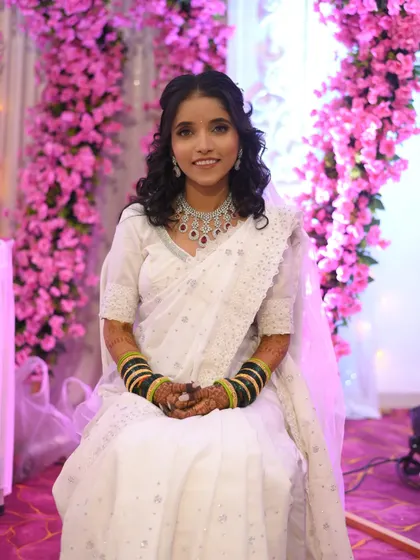 A smiling Buddhist bride against a backdrop of pink flowers. Her hair is styled with soft curls, adding a touch of modern romance to her traditional white saree.