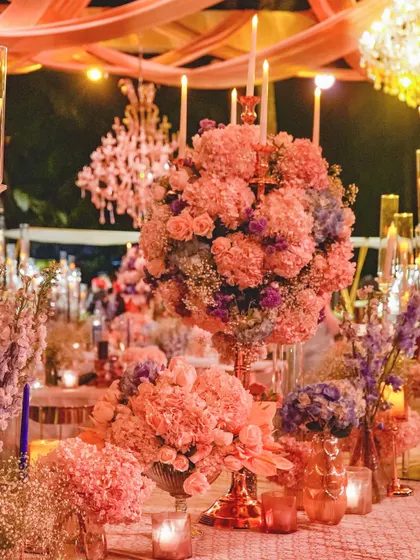 The entire tablescape is bathed in a warm, peachy light, enhancing the romantic feel of the pink and blue floral arrangements. This image captures the magical, fairytale quality of the event design.