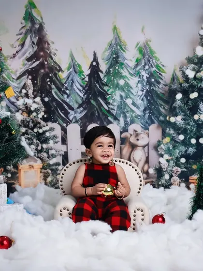 A joyful toddler enjoys a Christmas-themed photoshoot. Dressed in festive plaid, the child sits in a winter wonderland scene complete with decorated trees, snow, and presents.