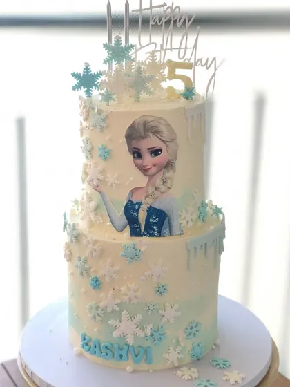 A two-tiered Frozen cake featuring an edible image of Elsa, surrounded by a flurry of buttercream and fondant snowflakes.