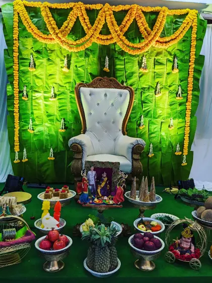 A full stage view of a Seemantham ceremony with a banana leaf backdrop. The setup includes a white throne, traditional doll arrangements, and a variety of decorated offering plates.