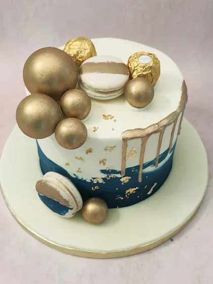 We design a cake of pure modern luxury with this blue and gold drip cake. A deep blue ombre effect is accented with gold drips, gold leaf, and an arrangement of golden spheres, Ferrero Rocher, and macarons, creating a sophisticated look for any celebration.
