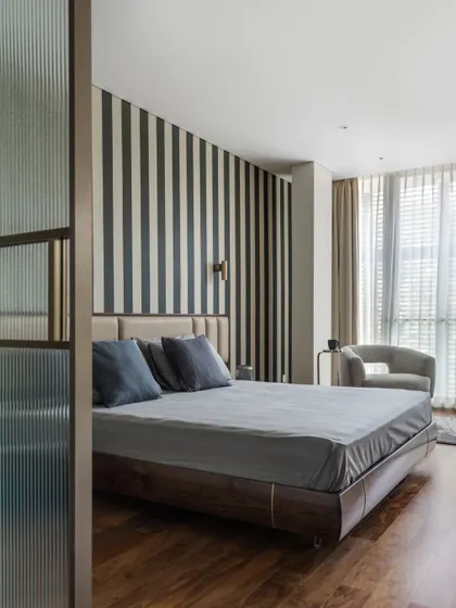 This bedroom is a study in refined comfort, featuring a bold striped wallpaper that adds graphic interest without overwhelming the space. A fluted glass partition provides a subtle separation, enhancing privacy while allowing light to filter through, creating a serene and layered retreat.