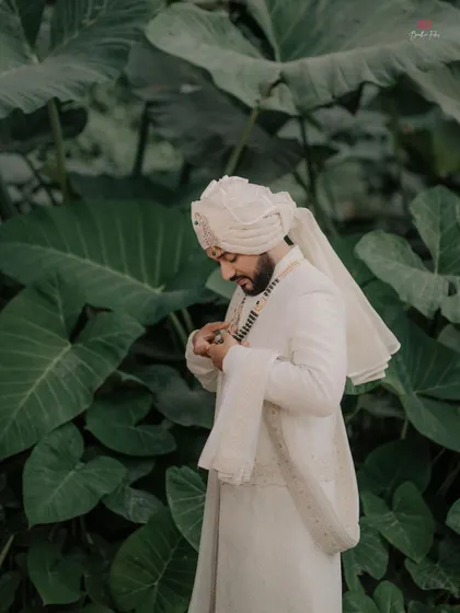 A handsome portrait of the groom in his white sherwani, set against a lush green backdrop.