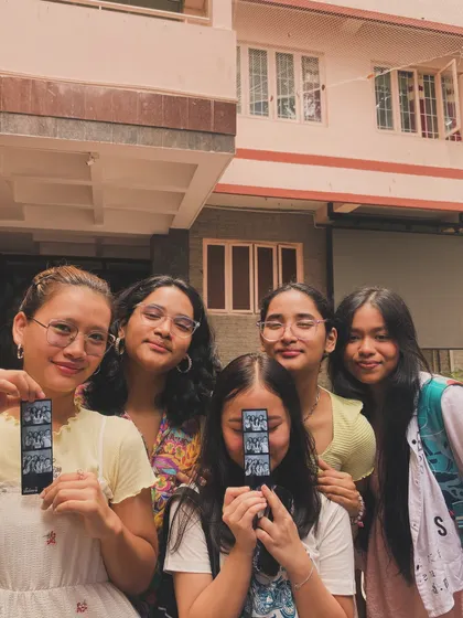 Five friends, some peeking from behind their photo strips, enjoying a sunny day at their college fest.