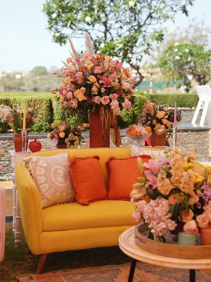 A bright yellow sofa adorned with orange and white cushions, surrounded by large, beautiful arrangements of pastel-colored flowers.
