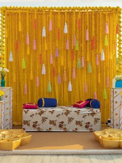 A beautiful rooftop Haldi setup. The marigold wall is accented with colorful tassels, and the traditional printed sofa completes the look.