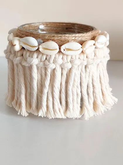 The natural off-white macrame jar is wrapped with jute and decorated with cowrie shells for a bohemian, beachy vibe.