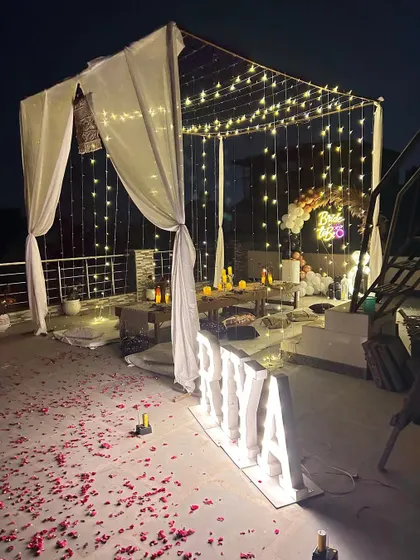 A romantic "Bride to Be" terrace decoration. The setup includes a canopy with white drapes and fairy lights, scattered rose petals, and large marquee letters for the name.